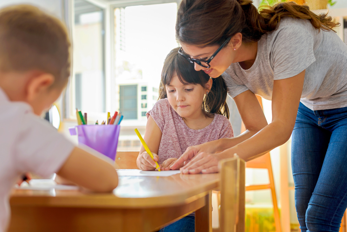 Children drawing with smiling preschool teacher assisting them